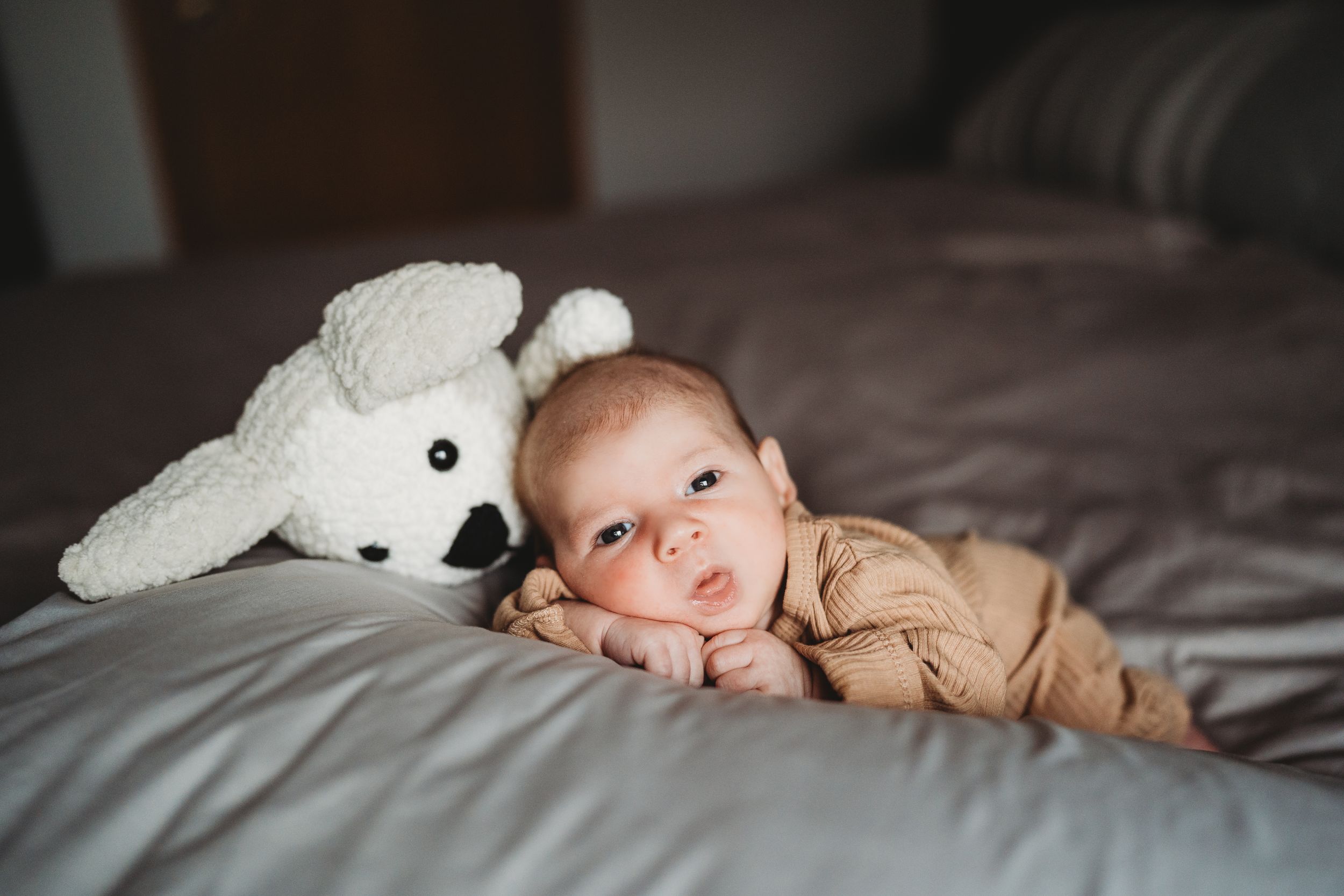Baby in beige outfit lying on bed next to white plush bunny toy.
