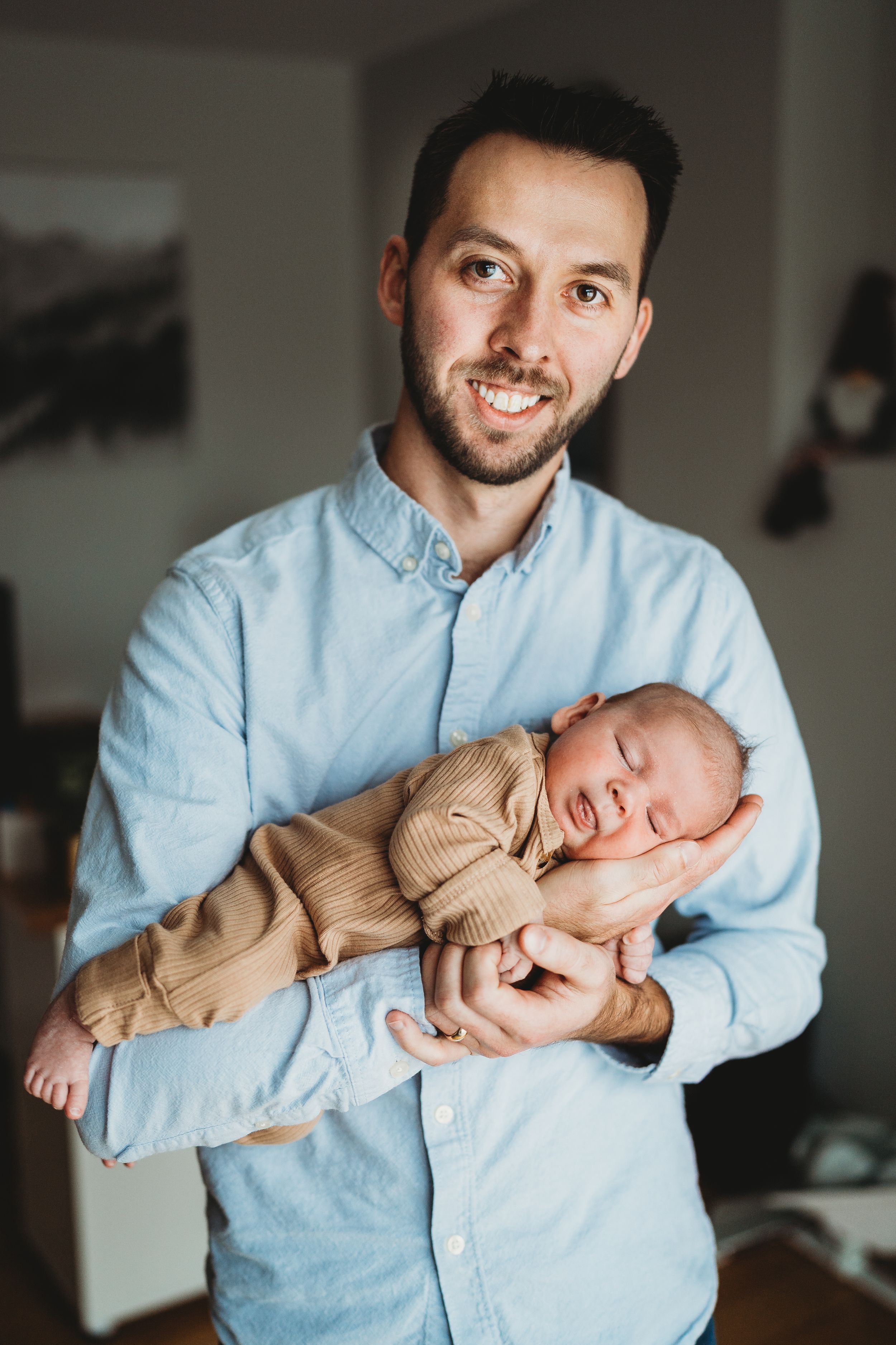 Father in light blue shirt holding sleeping newborn in tan outfit.