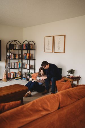 A couple shares a tender moment on their brown couch near a bookshelf with framed art.