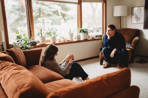 Two people relax on a brown couch in a living room with large windows and plants.