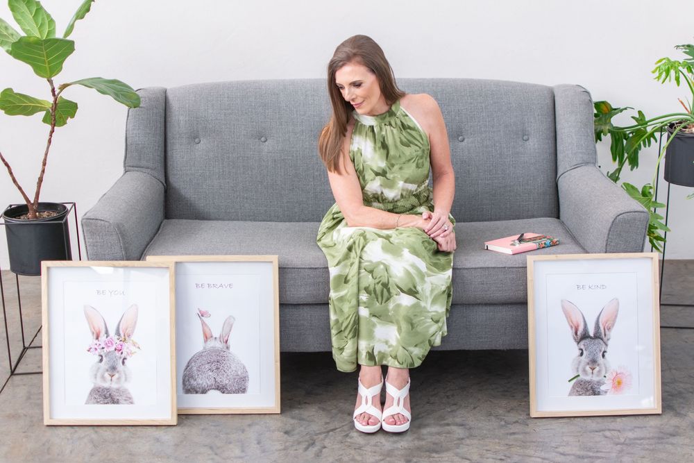 Woman in green floral dress sitting on gray sofa surrounded by framed bunny rabbit art prints on floor.