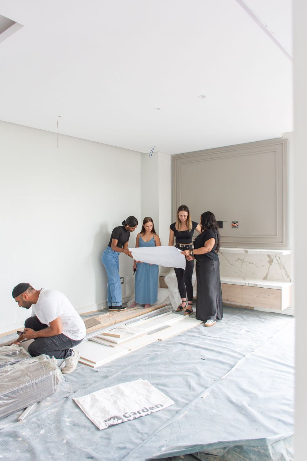 Group reviewing blueprints in a room under renovation with marble feature wall and flooring samples on protective sheeting.