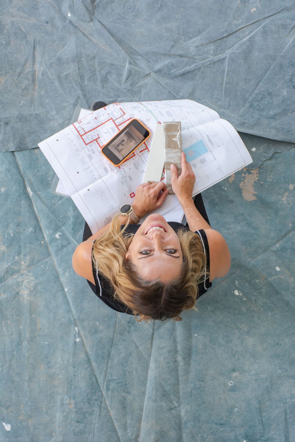 Overhead view of a smiling woman reviewing blueprints and holding paint swatches on a drop cloth floor.