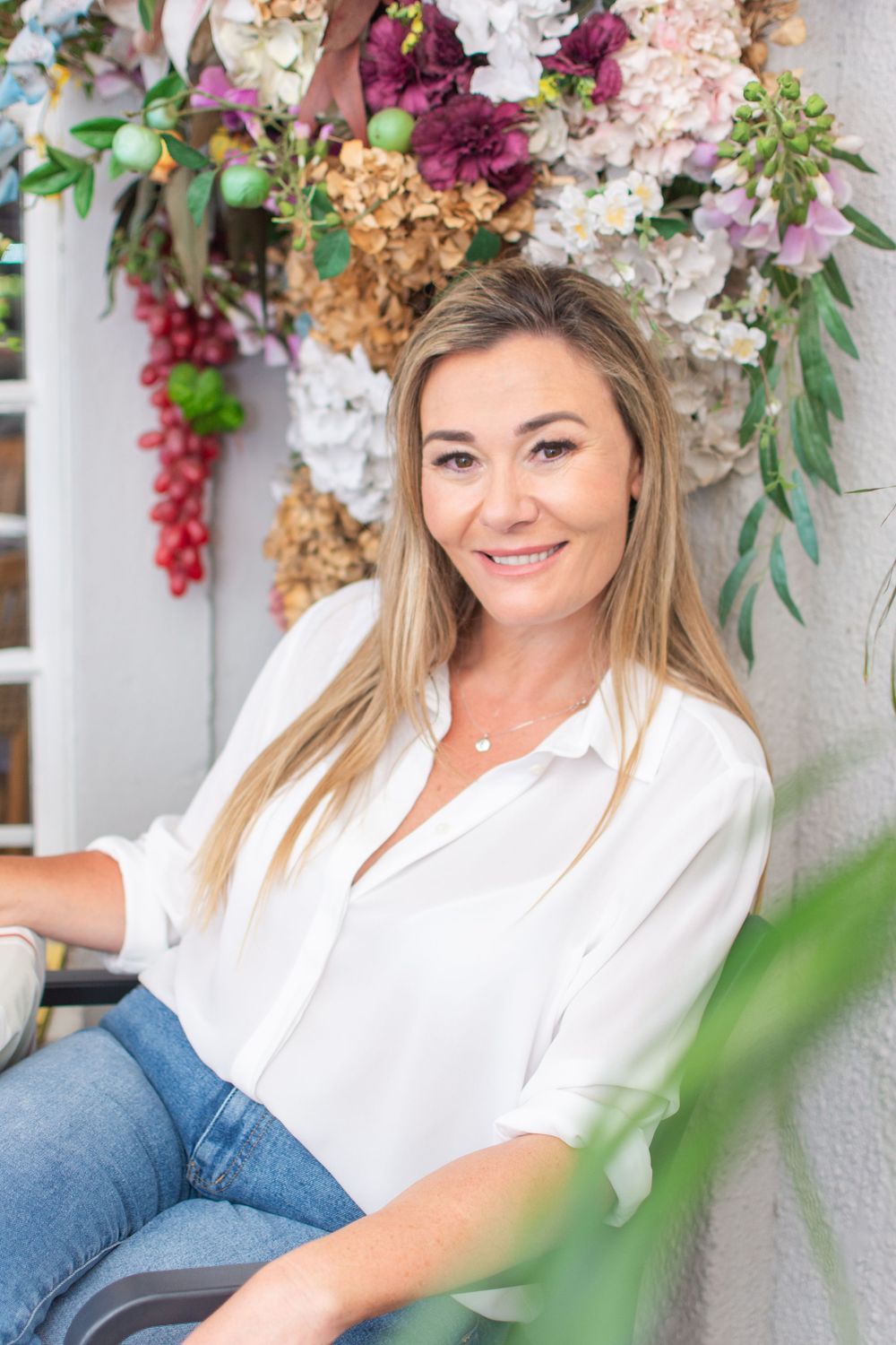 Smiling blonde woman in white blouse and jeans sitting relaxed against a wall with colorful floral arrangement backdrop.