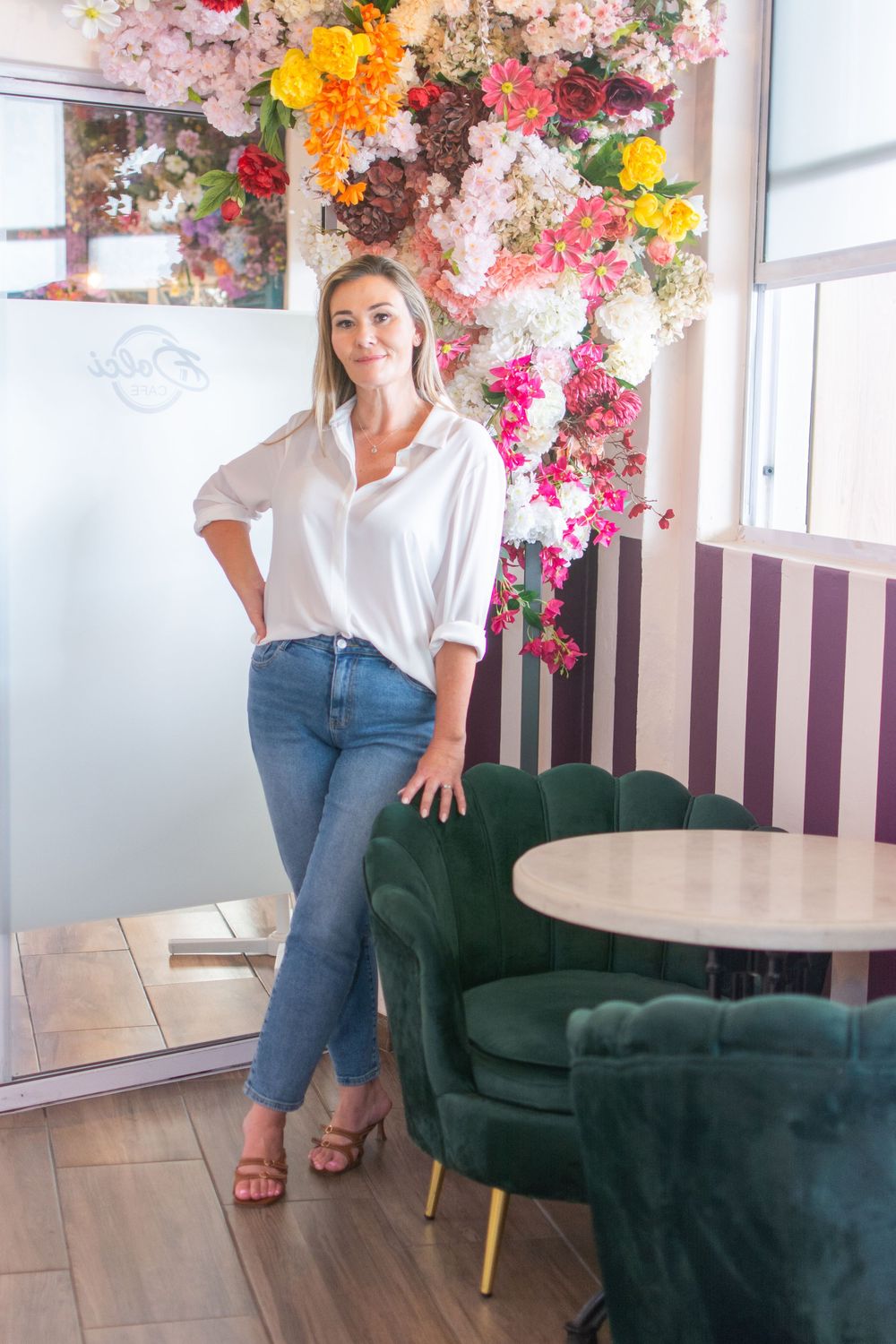 Woman in white shirt and jeans poses in a floral cafe with green velvet chairs and striped walls.