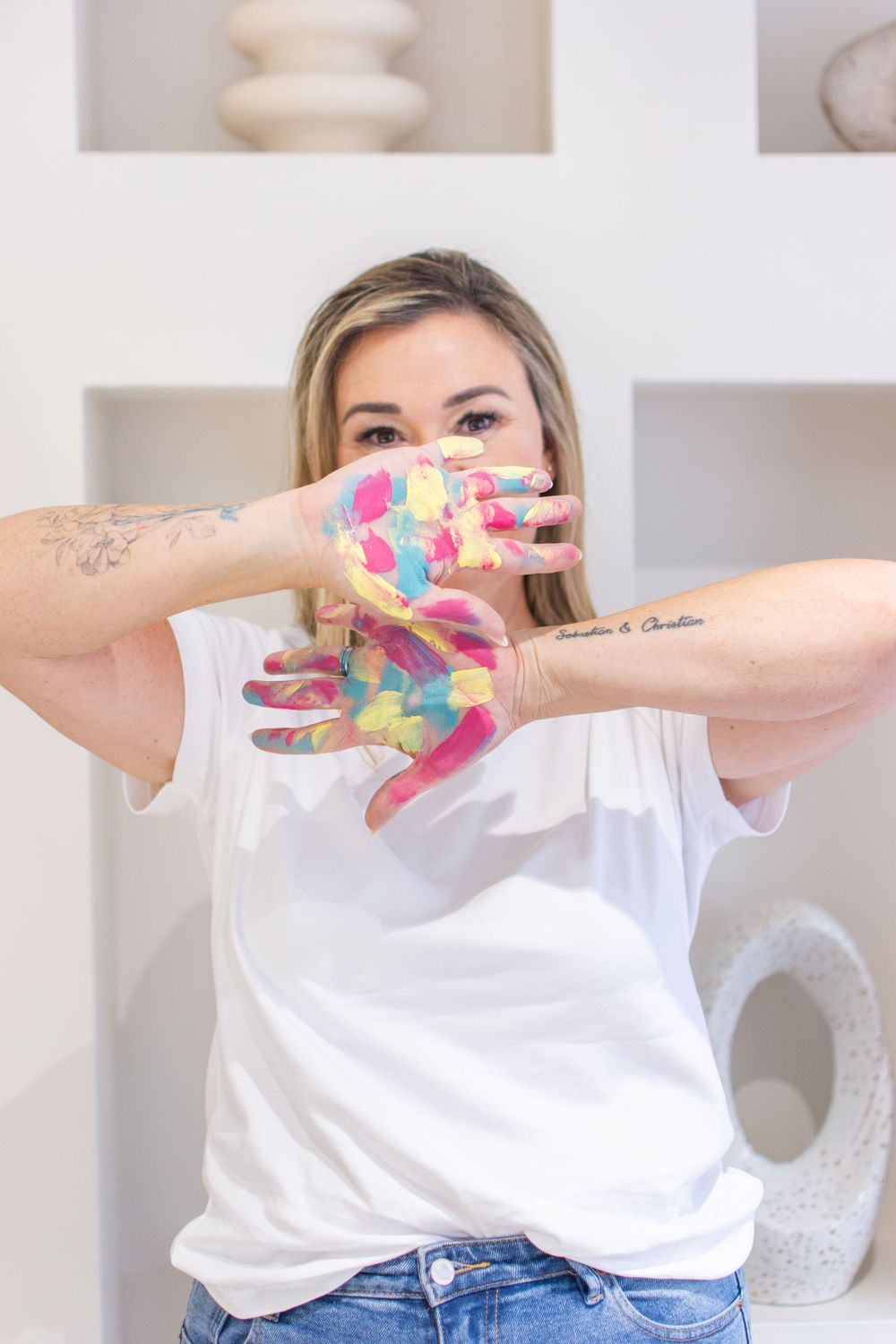 Woman in white t-shirt showing colorful paint-covered hands, with tattoos visible, standing in front of white shelves.