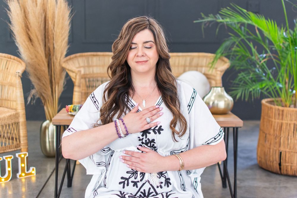 Woman in white kimono meditating with hands on heart and belly, seated outdoors in a boho-styled space.