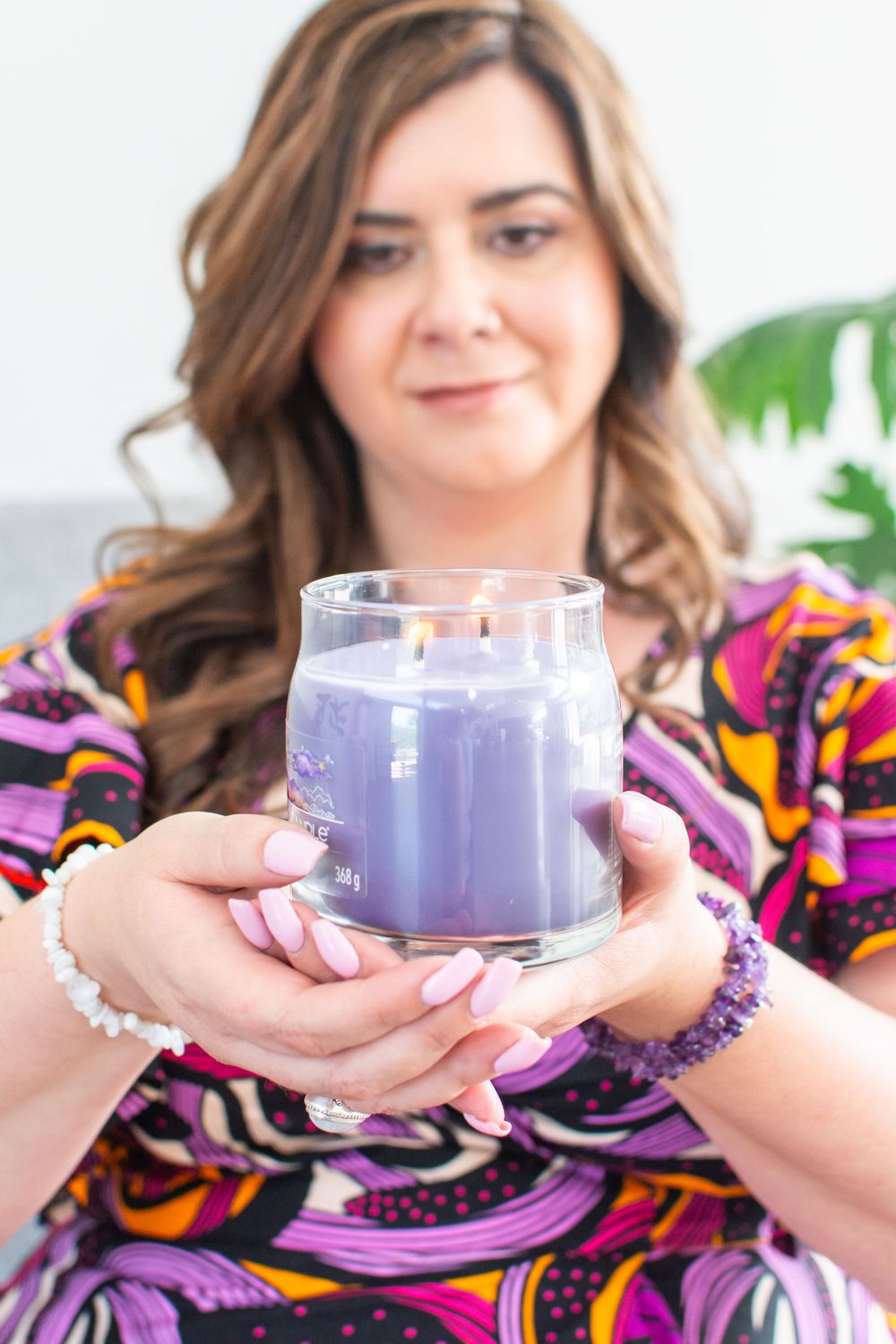 Woman in colorful dress holding a lit purple lavender jar candle in a glass vessel with both hands.