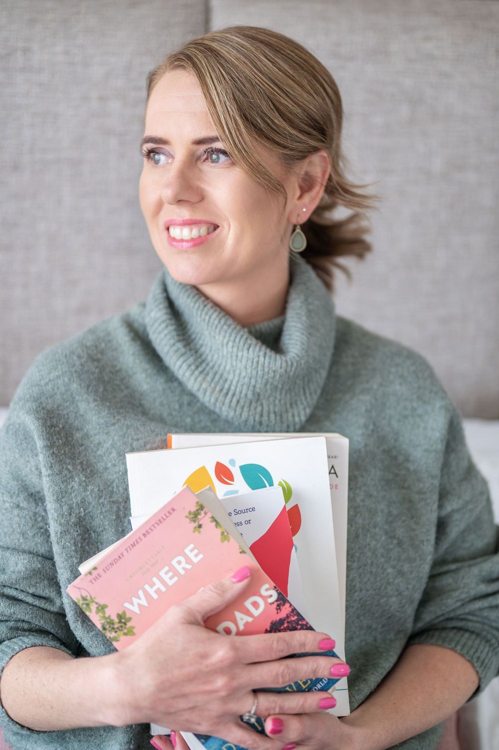 Smiling person in sage green turtleneck sweater holding colorful books against grey background.