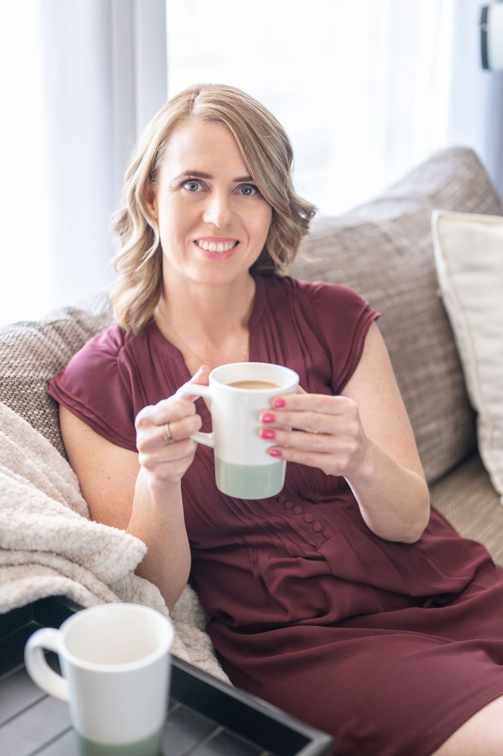 Person in burgundy dress relaxes on gray couch with white coffee mug while enjoying a cozy morning at home.