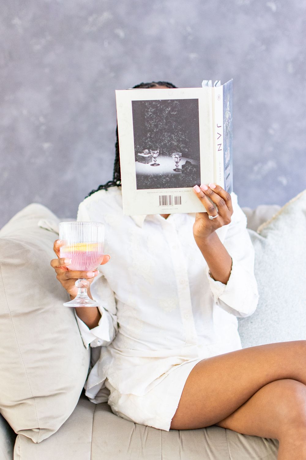 A woman sitting on a white couch holds up a small book against a grey textured background.