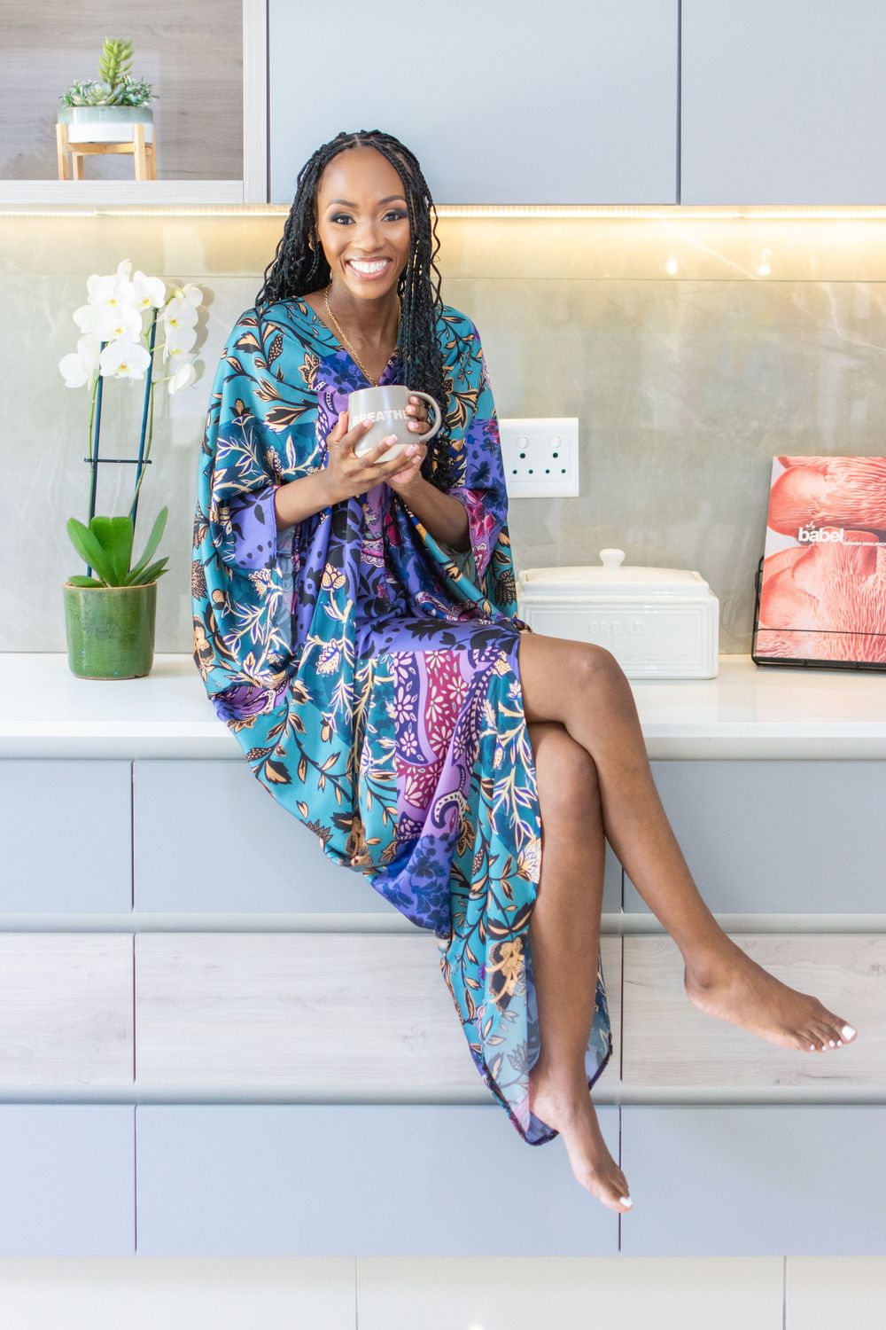 Person in vibrant floral kimono robe relaxes on modern white bench with orchid decor and coffee cup.