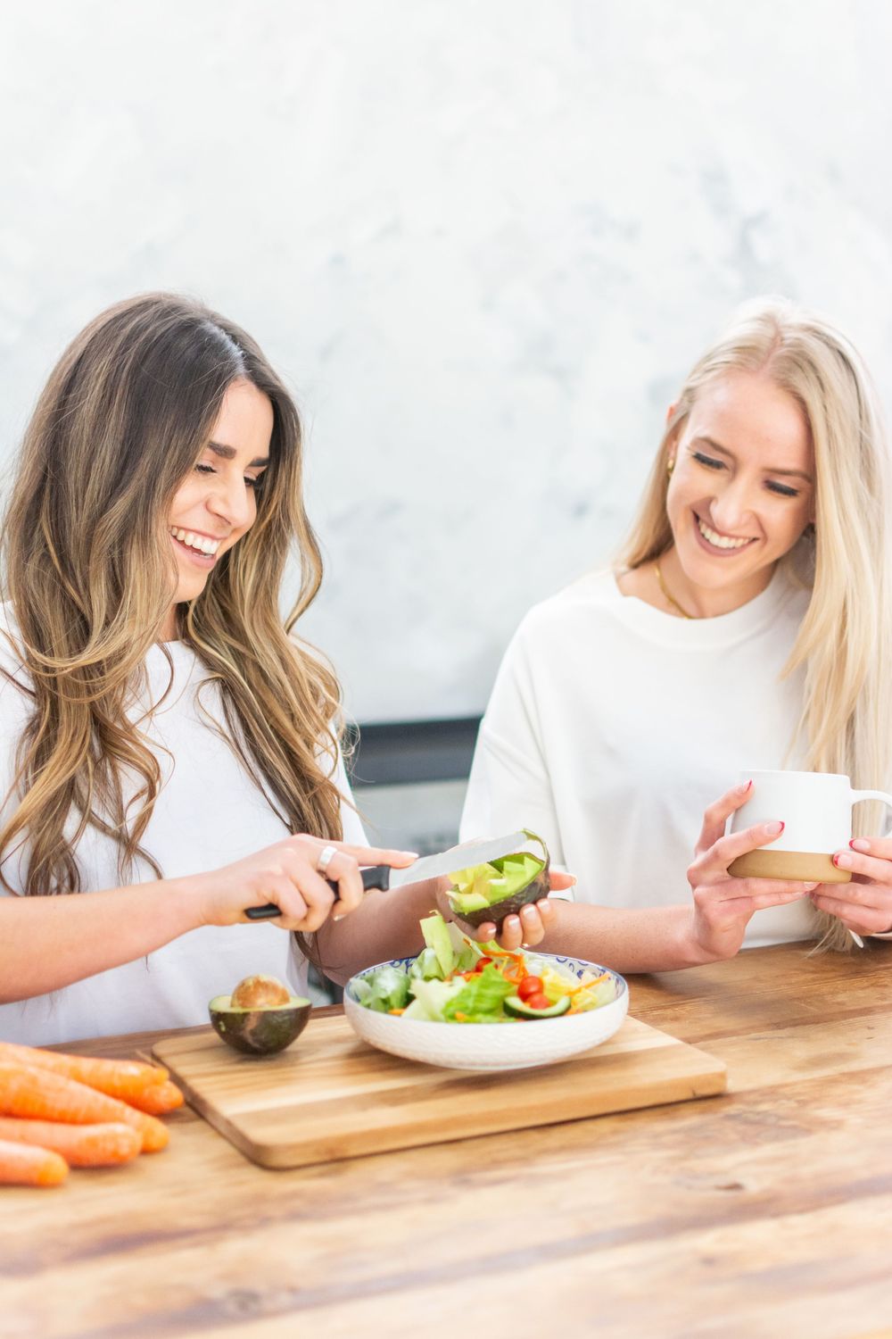 Two women in white shirts preparing healthy food and smiling while cutting vegetables at wooden table.
