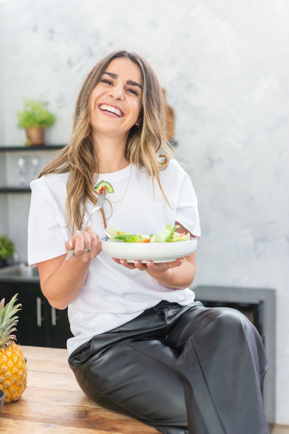 Woman in a white t-shirt and black pants enjoys a healthy meal while sitting at a wooden table with fresh pineapple.