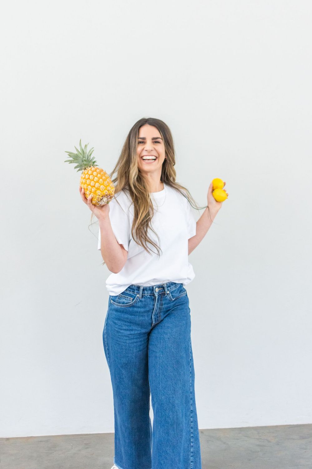 Woman in a white t-shirt and wide-leg jeans holds a fresh pineapple and yellow bell pepper against a white wall.