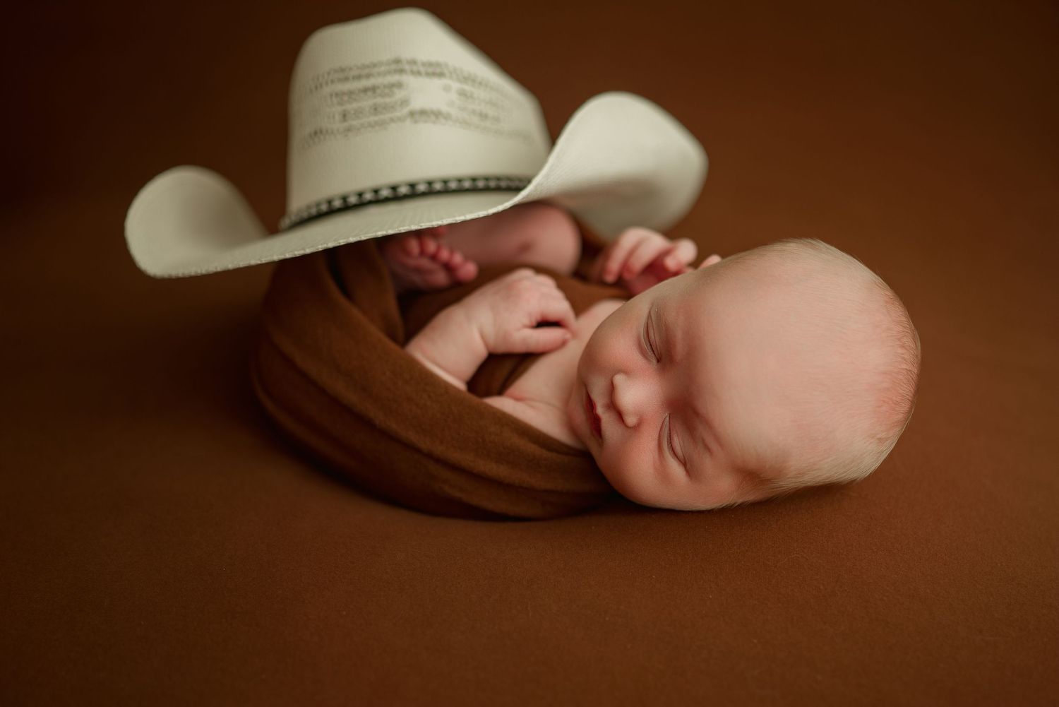 Newborn baby boy with cowboy hat wrapped in brown swaddle during studio newborn session in Clarksville, Tennessee.