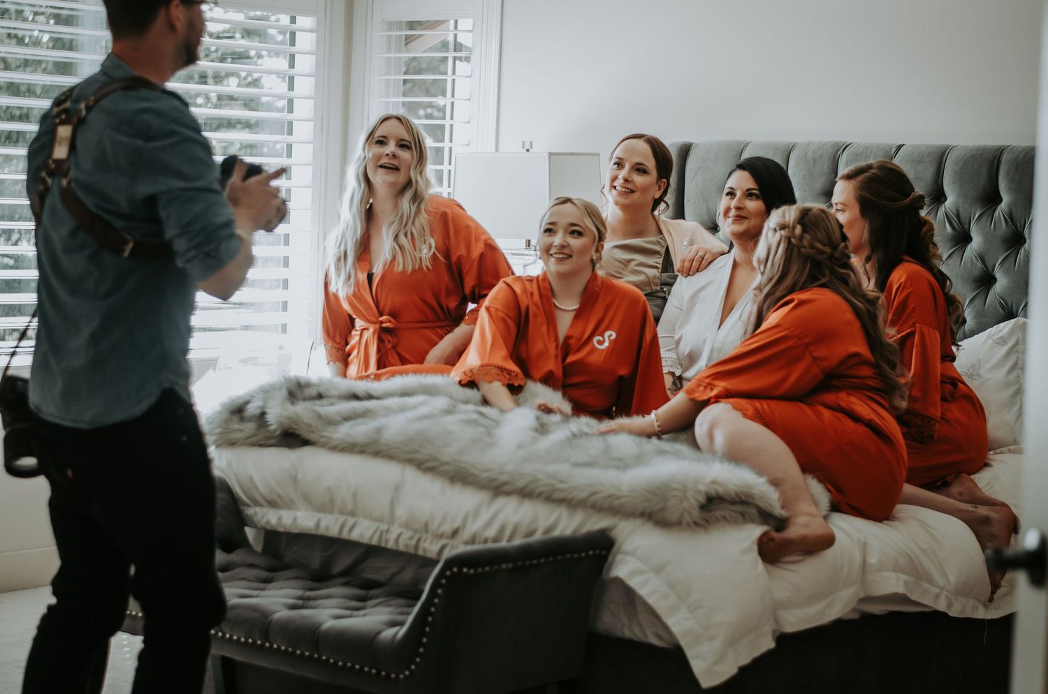 Bridesmaids in orange robes laughing on bed while photographer captures getting-ready wedding moments.