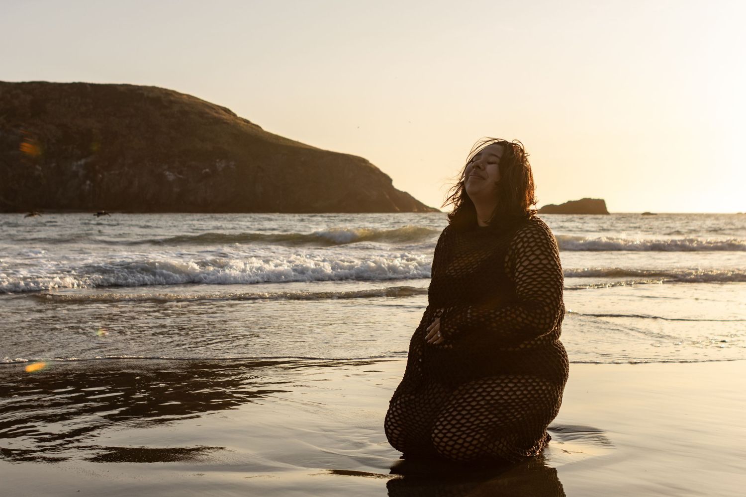 An expecting mama sits on the beach in all black, wearing a black two piece, and a long sleeve crochet coverup