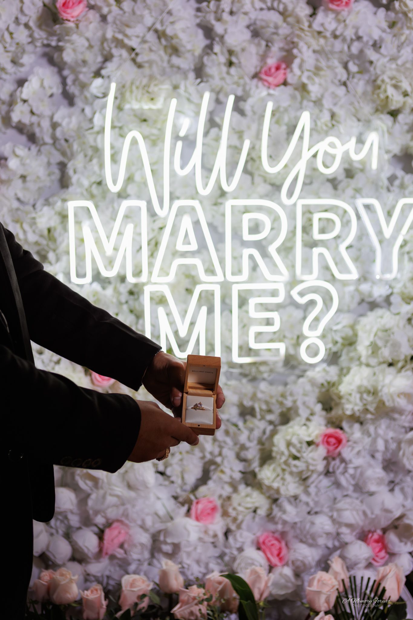 Person holding an open ring box in front of a floral wall with a neon "Will You Marry Me?" sign.