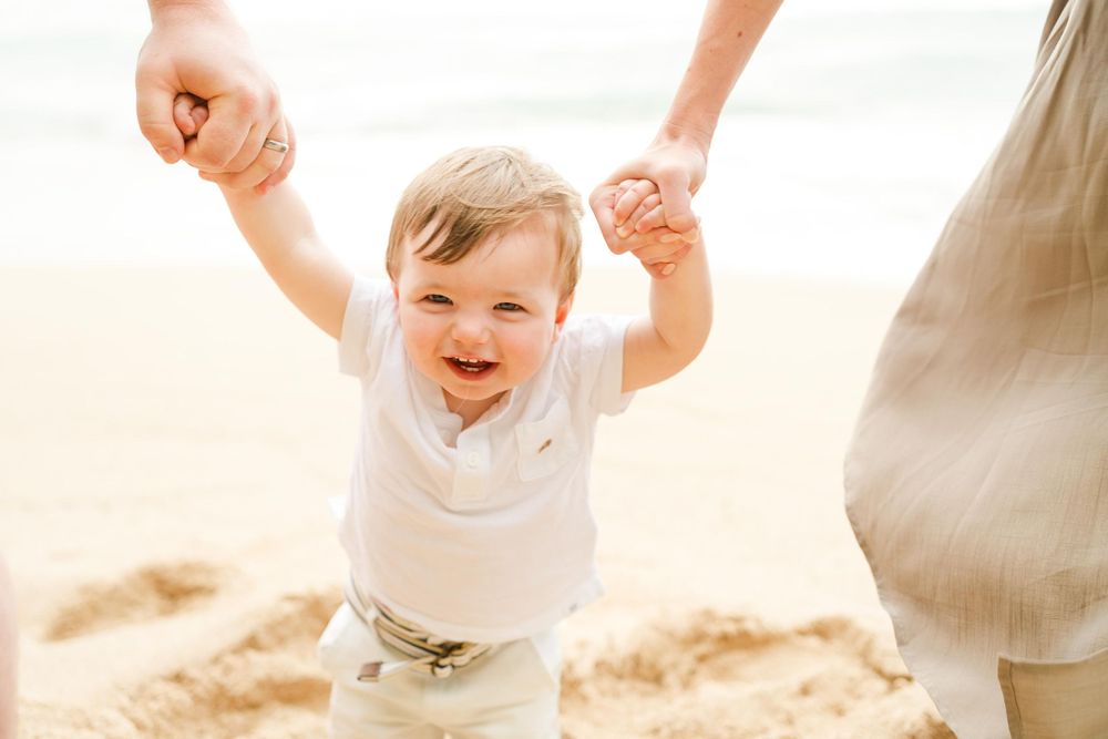 A joyful small child in white clothing walks along the beach while being supported by adult hands. Oahu Family Photographer