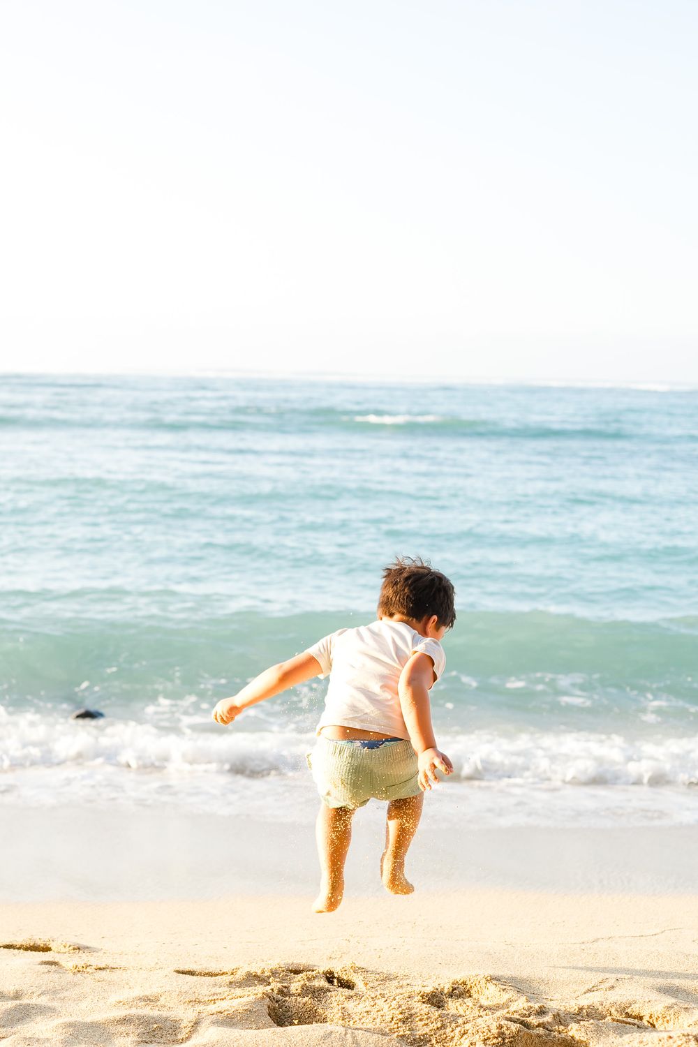 A small child jumps joyfully on a sunny beach with turquoise ocean waves in the background.