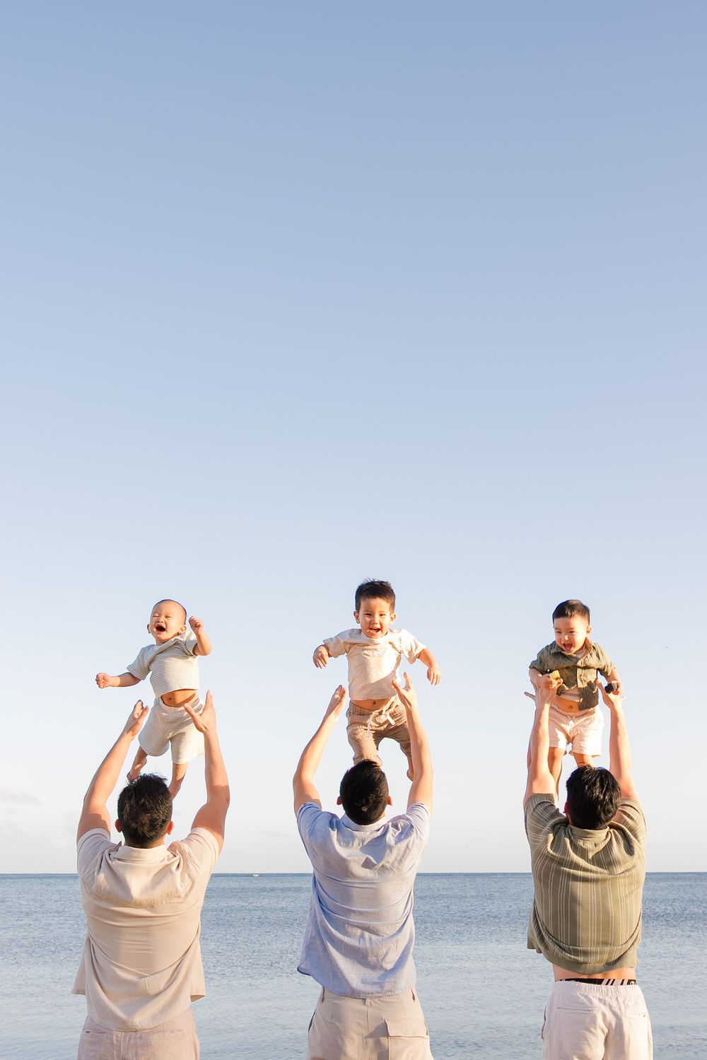 Three adults lift small children up against a blue sky and ocean backdrop at the beach.