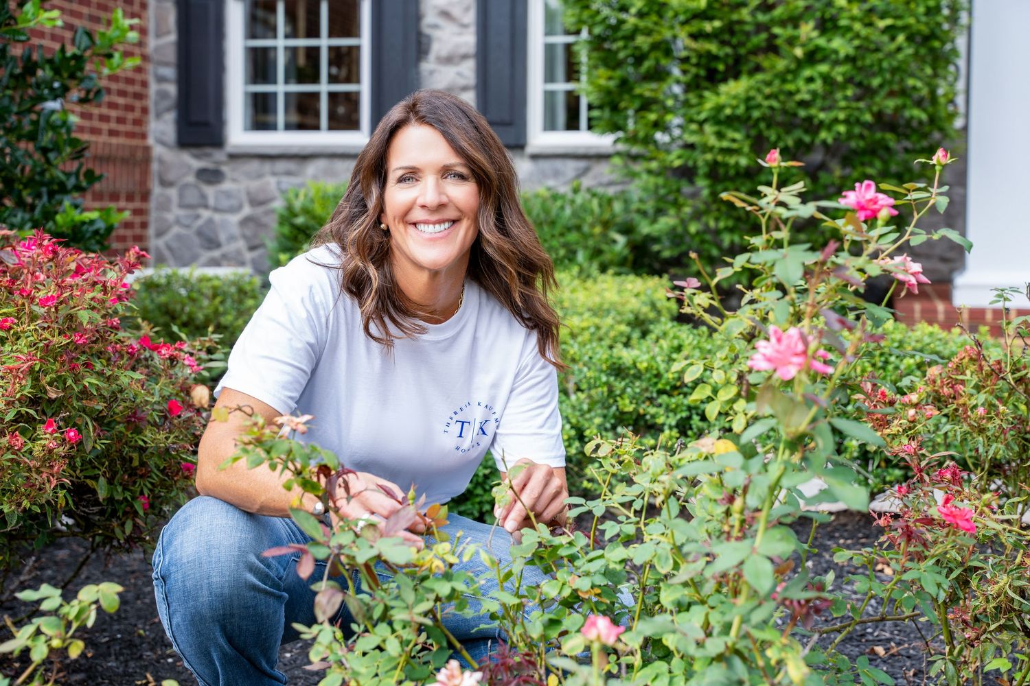 Person gardening and tending to blooming flowers outside a brick house with white windows.