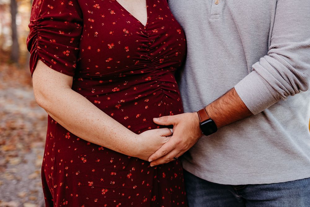 A couple standing each other and they are both holding each others hand on the expectant mothers belly.