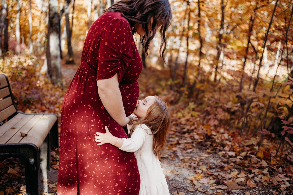 An expectant mother and her daughter is kissing her belly. The image is taken during fall, leaves are on the ground.