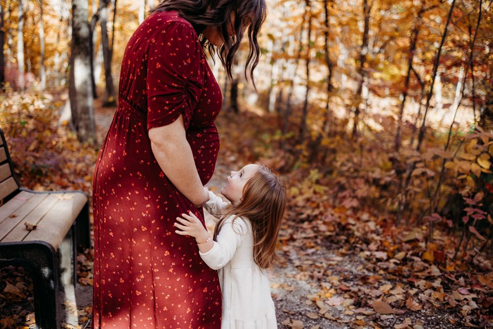 An expectant mother and her daughter smiling up at her on a trail in the forest. The image is taken during fall, leaves are on the ground.