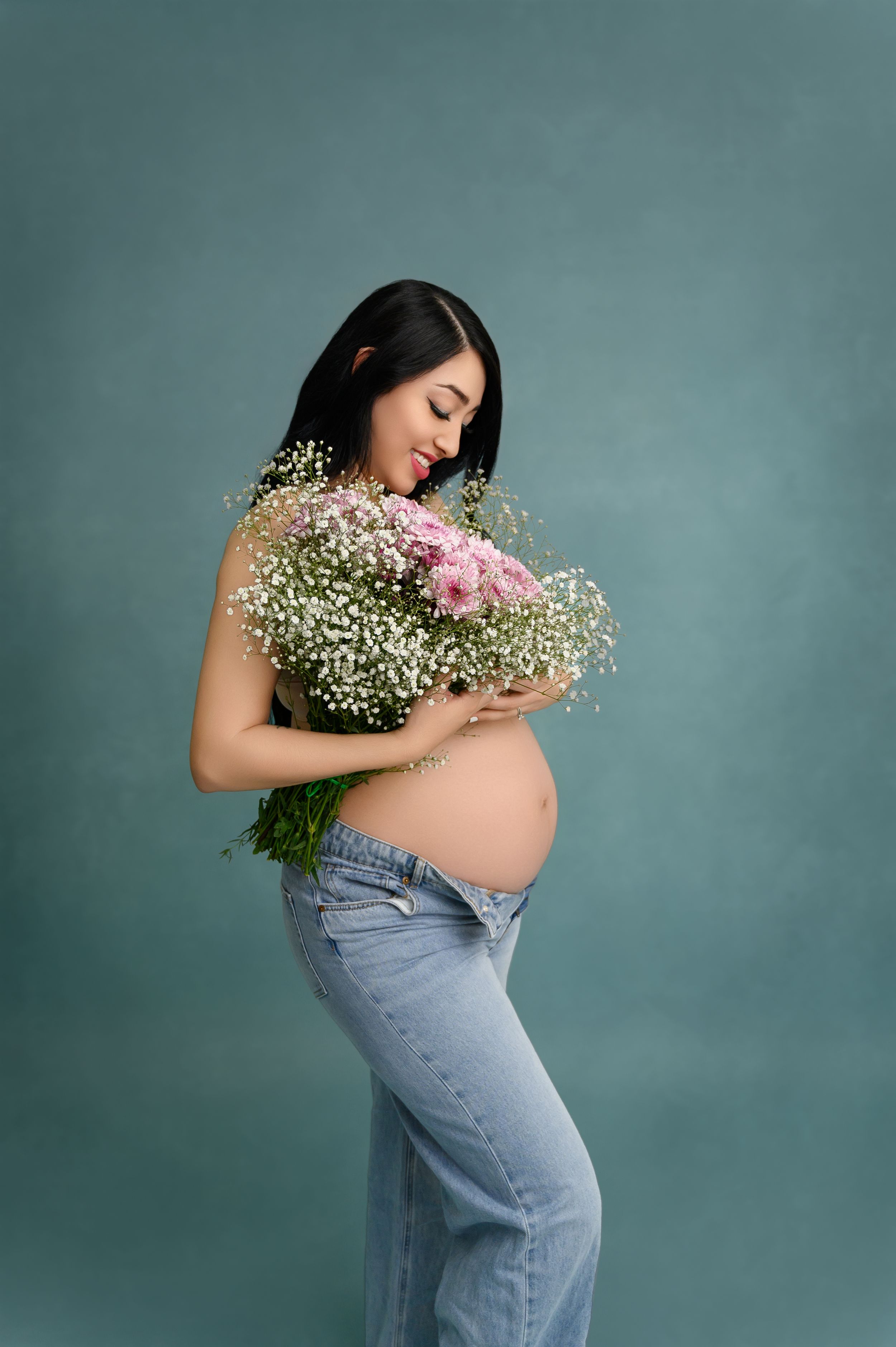 Pregnant woman in jeans holding baby's breath and pink flowers against teal background.