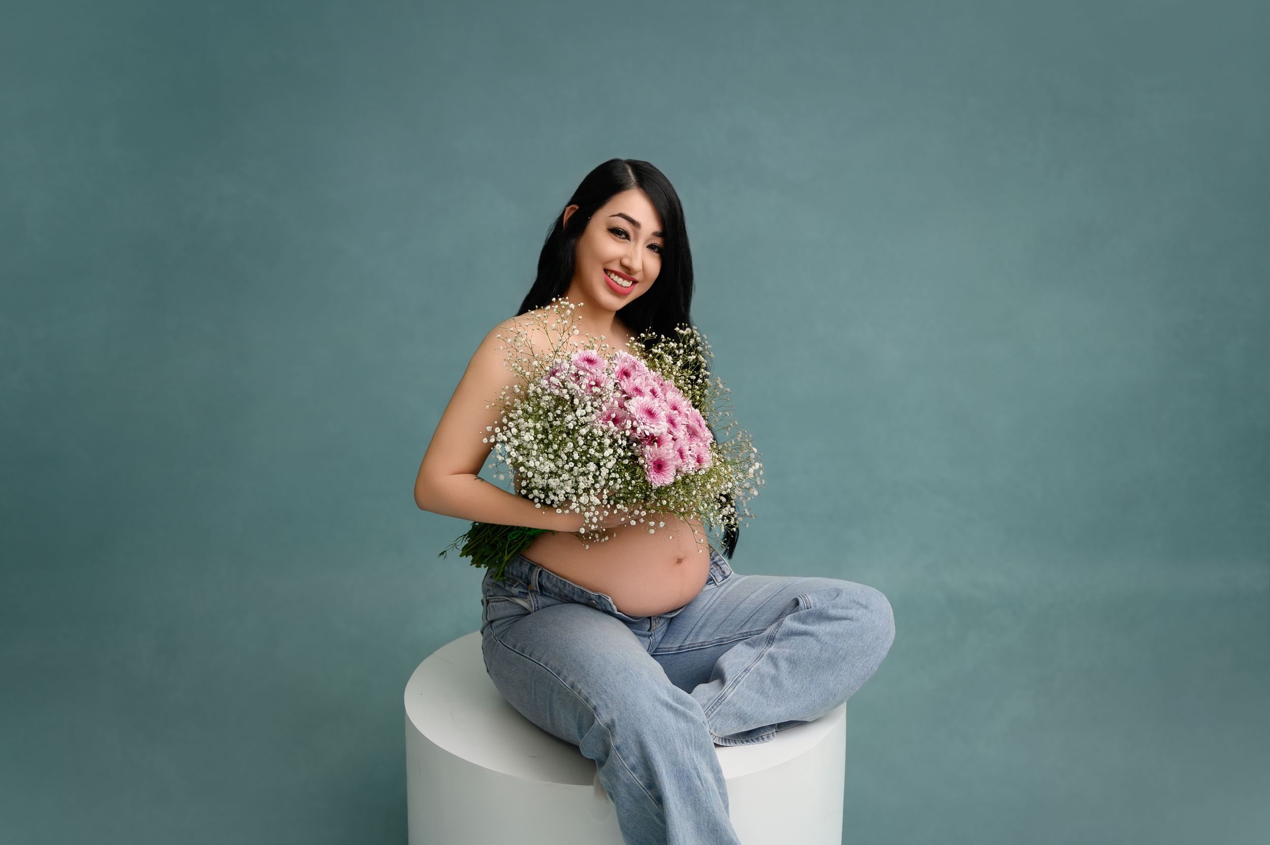 Expectant mother in denim poses with delicate flowers against turquoise studio backdrop.