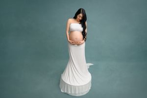 Maternity portrait series in flowing white dress against teal studio background.