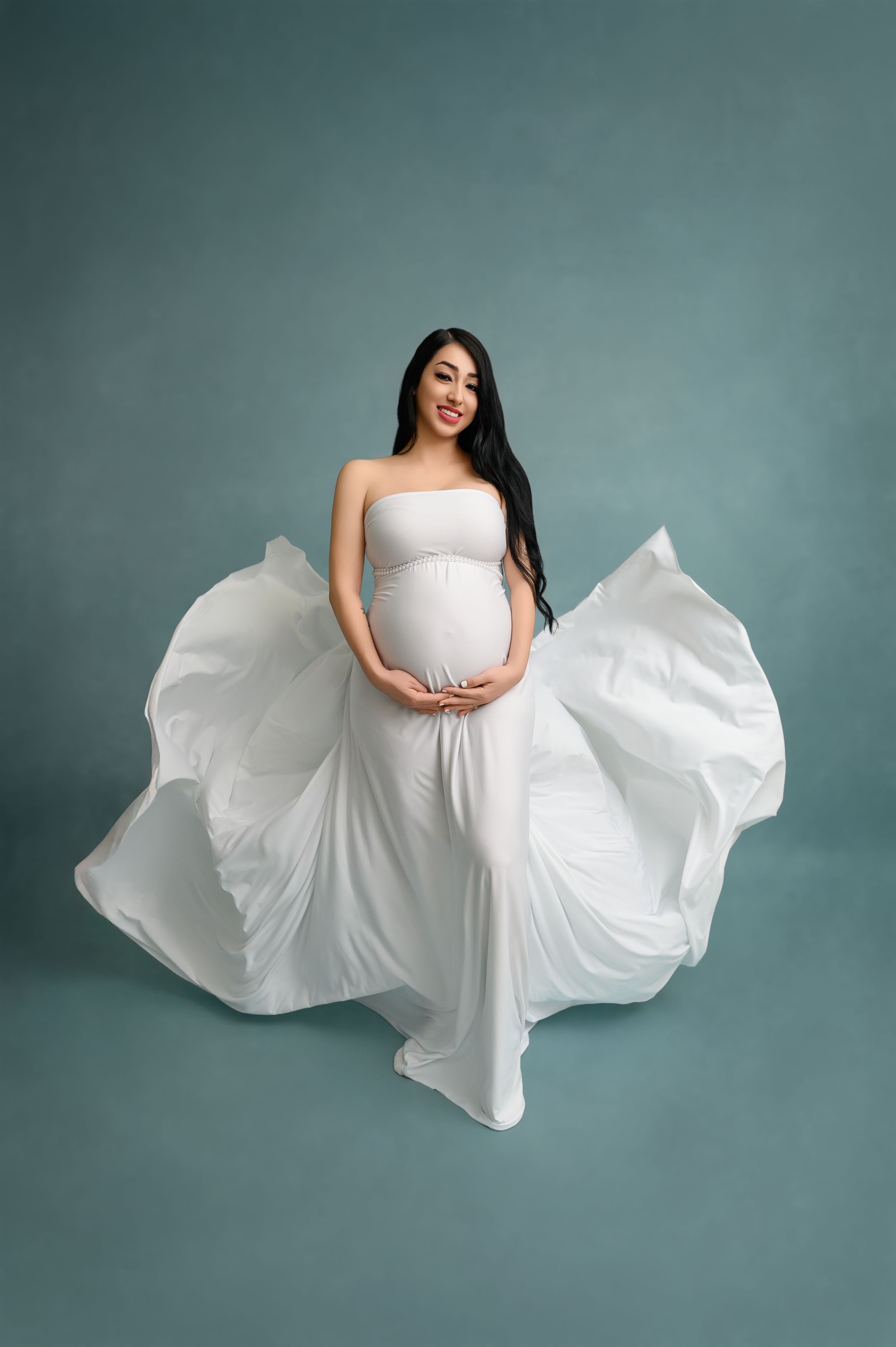 Dramatic maternity portrait with flowing white dress against teal background.