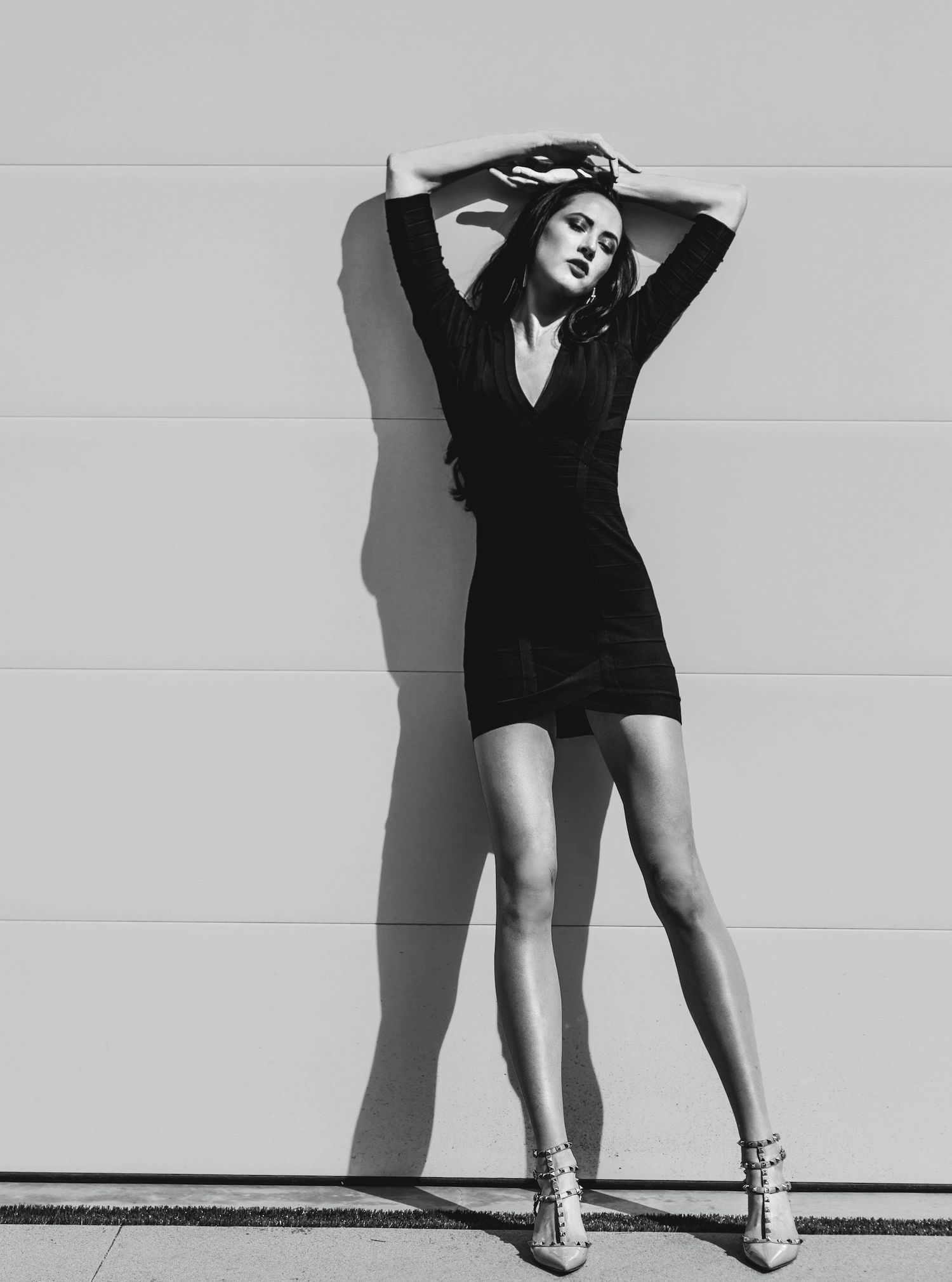 High-fashion Black and White image, long haired brunette woman with long legs, eyes closed, little black dress, sun & shadow leaning on wall