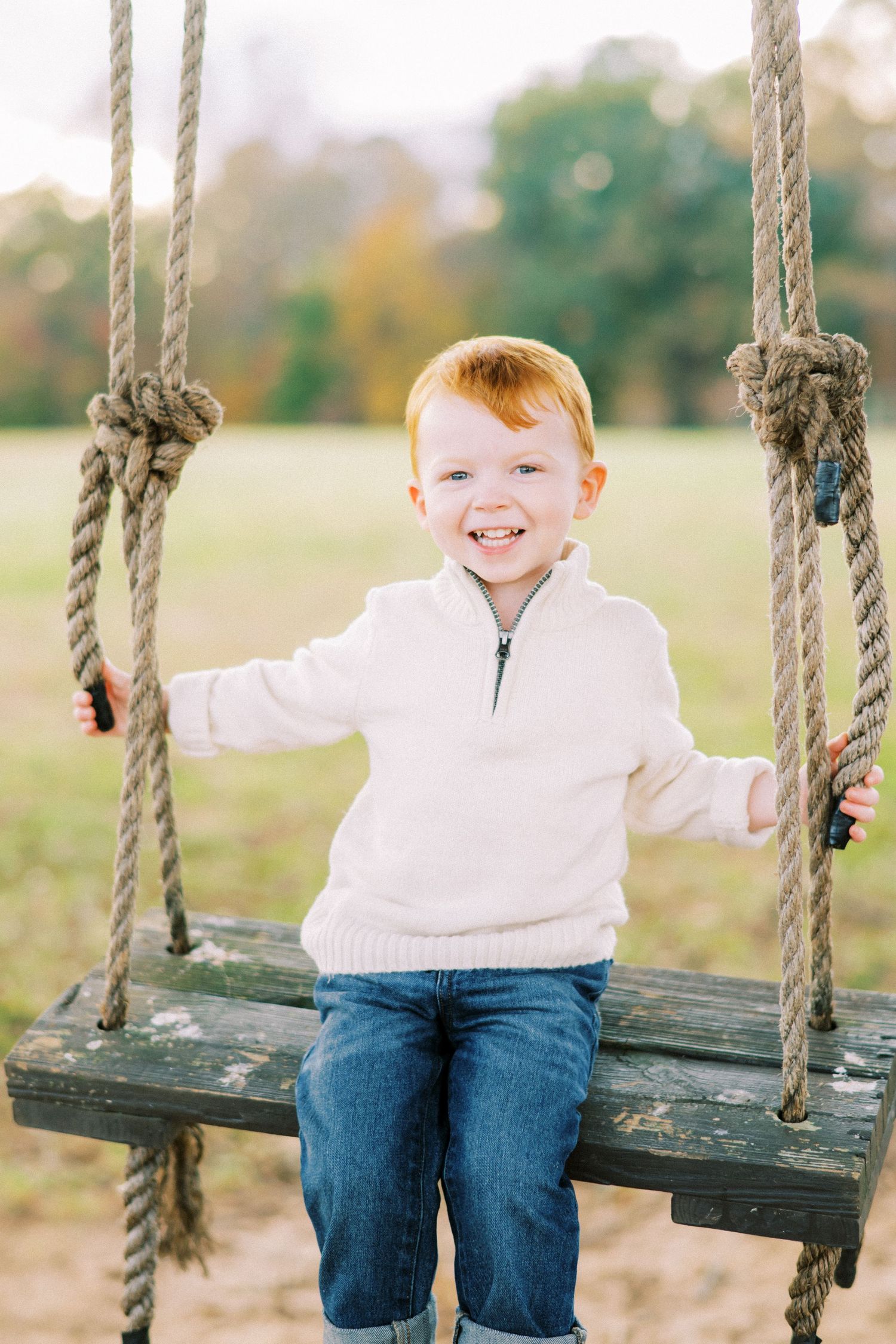 A child in a white sweater enjoys playing on a rustic rope swing outdoors.
