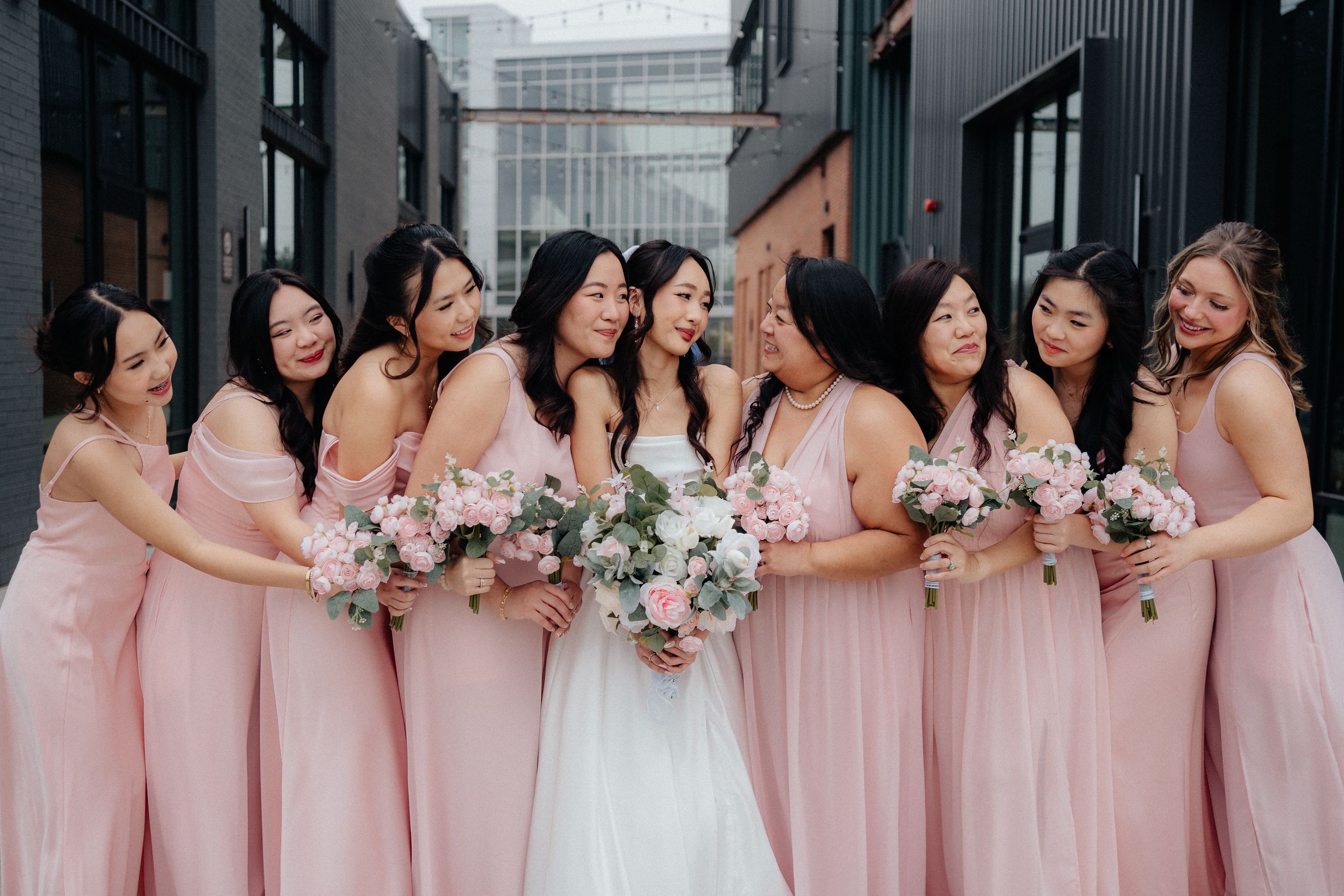 Bridesmaids in pink dresses and bride in white gown pose together in a long line holding floral bouquets.