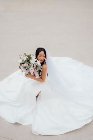 Bride in flowing white wedding gown poses gracefully on light colored floor with white floral bouquet.