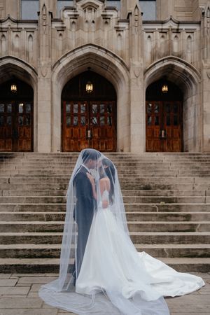 A wedding couple stands under Gothic arches on stone steps of a church, draped in a long flowing veil.
