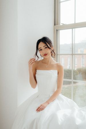 A bride in an elegant white ball gown poses gracefully near bright windows in a light-filled room.