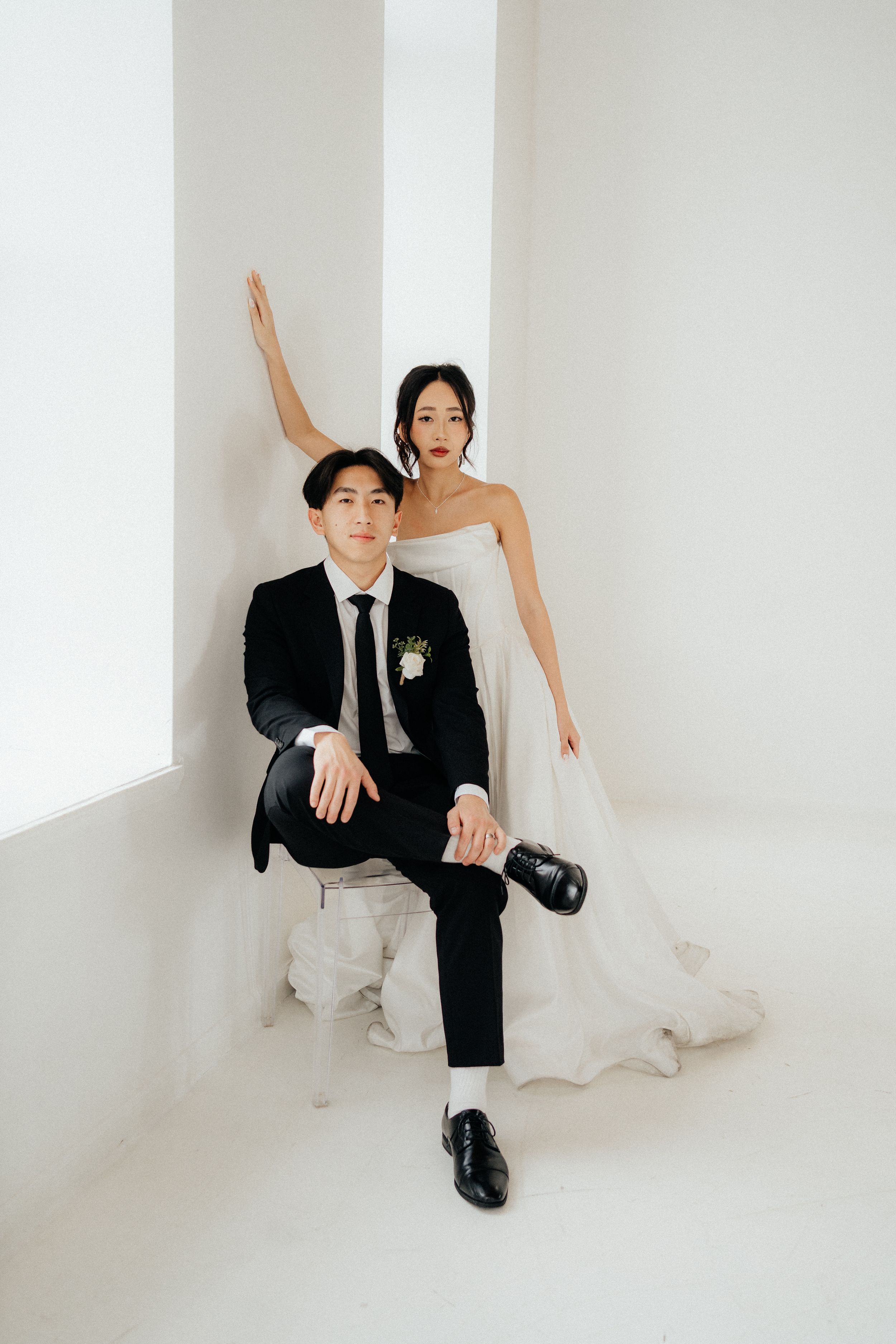 Wedding couple in black and white attire pose playfully against minimalist white studio backdrop.