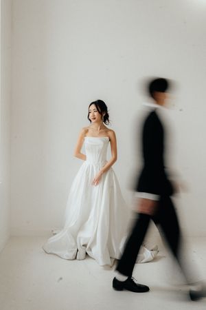 Artistic wedding photo with bride in white gown and blurred groom walking past in minimalist studio setting.
