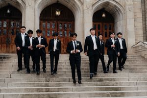 Groomsmen in black tuxedos stand on stone steps of a historic building for a formal wedding photo.
