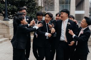 A group of groomsmen in black suits share a joyful moment laughing together outside a building.