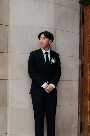 Three groomsmen in black suits and white boutonnieres lean against a light-colored stone wall at a formal event.