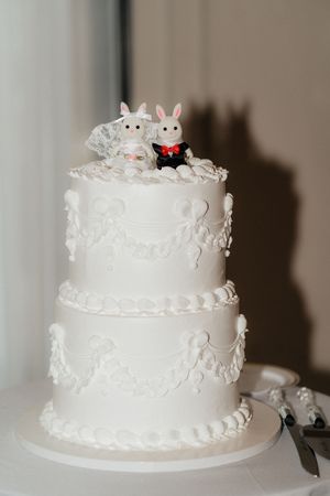 White tiered wedding cake decorated with delicate lace patterns and topped with bunny figurines.