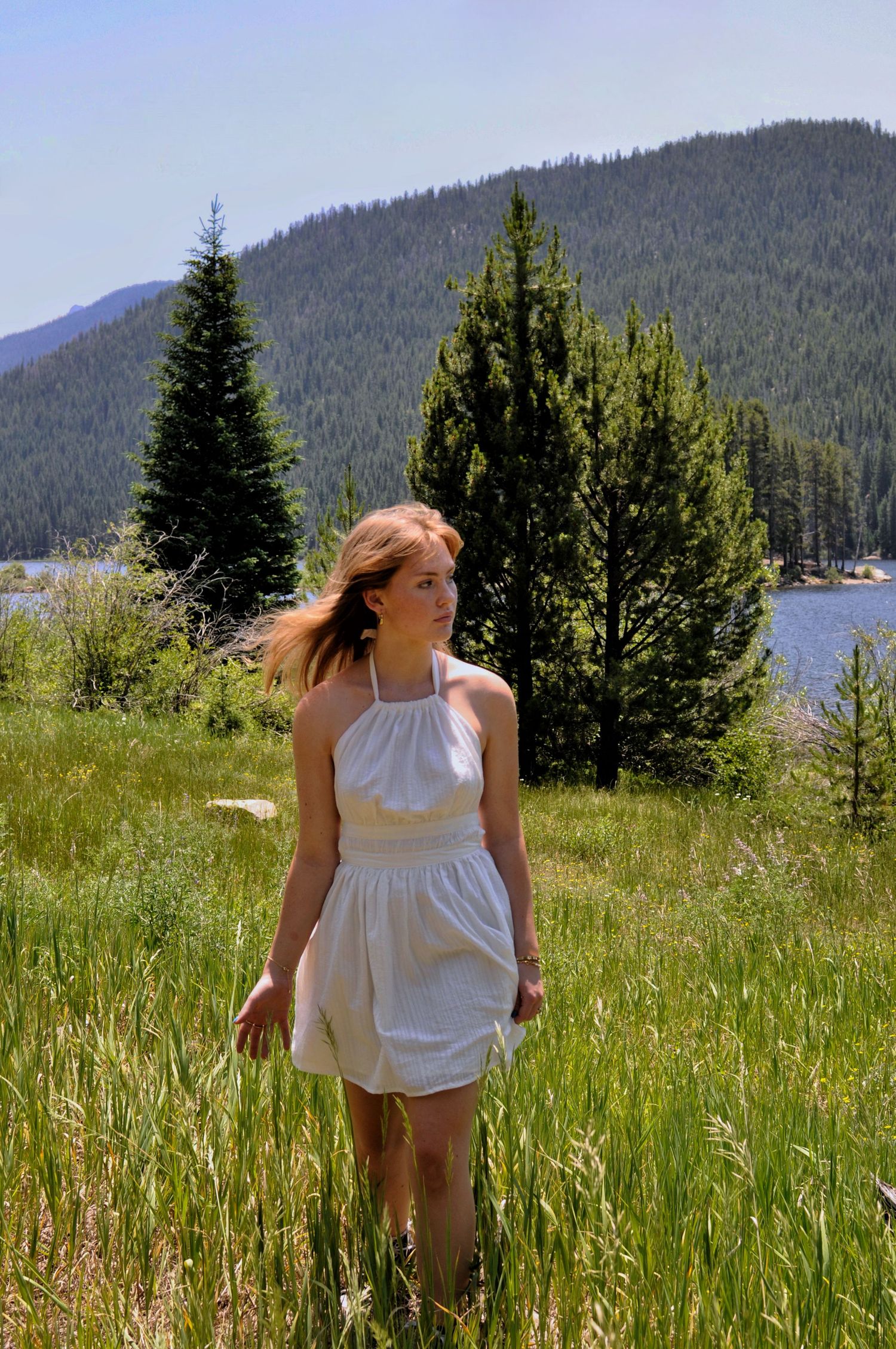 Young lady walks through a grassy field, with a mountain lake scene behind.
