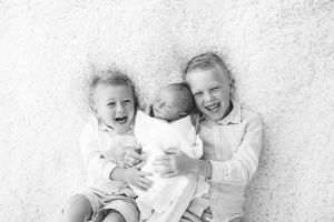 Black and white photo of two laughing siblings holding their newborn baby sibling.