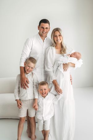 Happy family of five in all-white outfits with newborn baby posing in a bright studio.