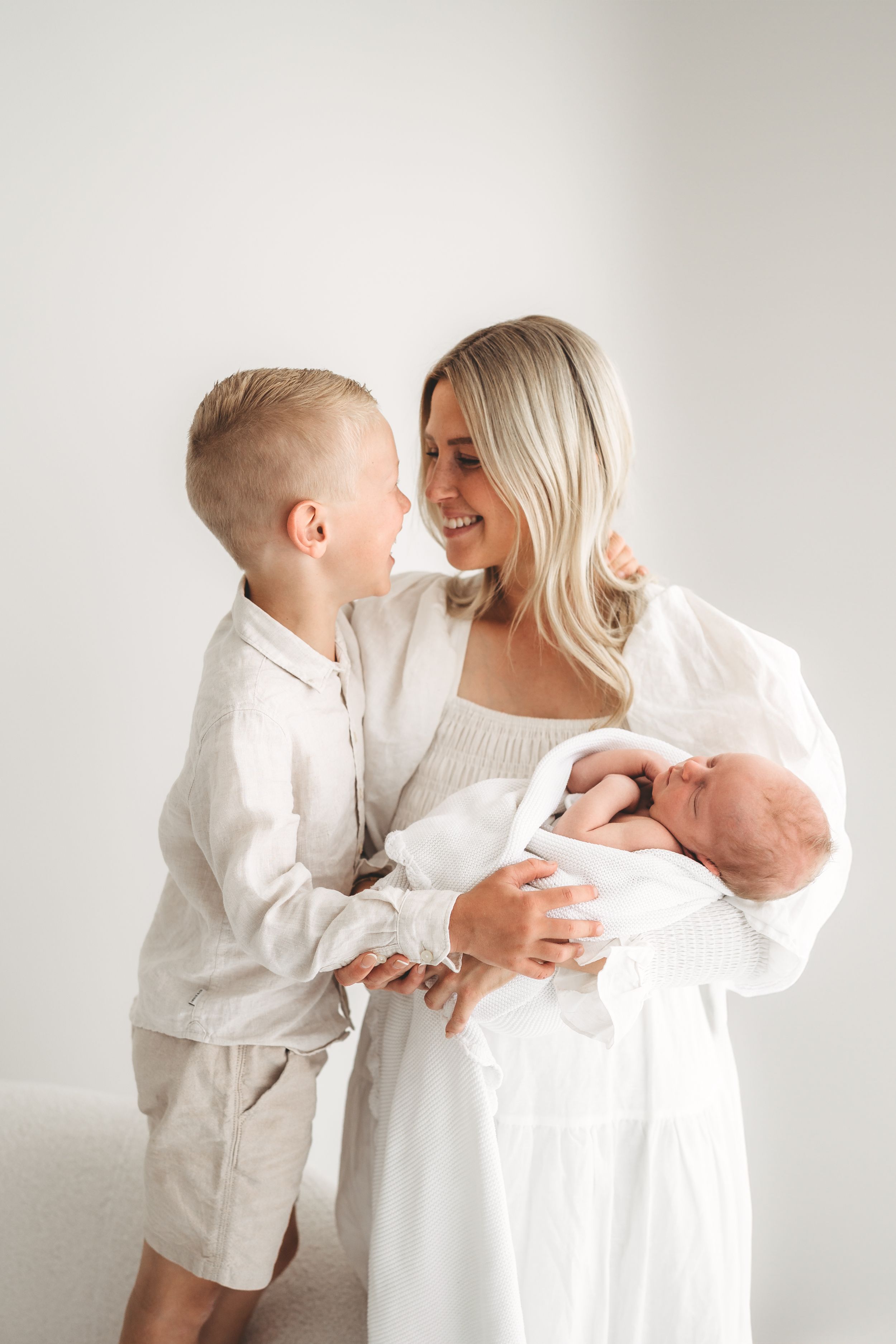 Smiling mother in white dress holding newborn baby while young son leans in to kiss her.